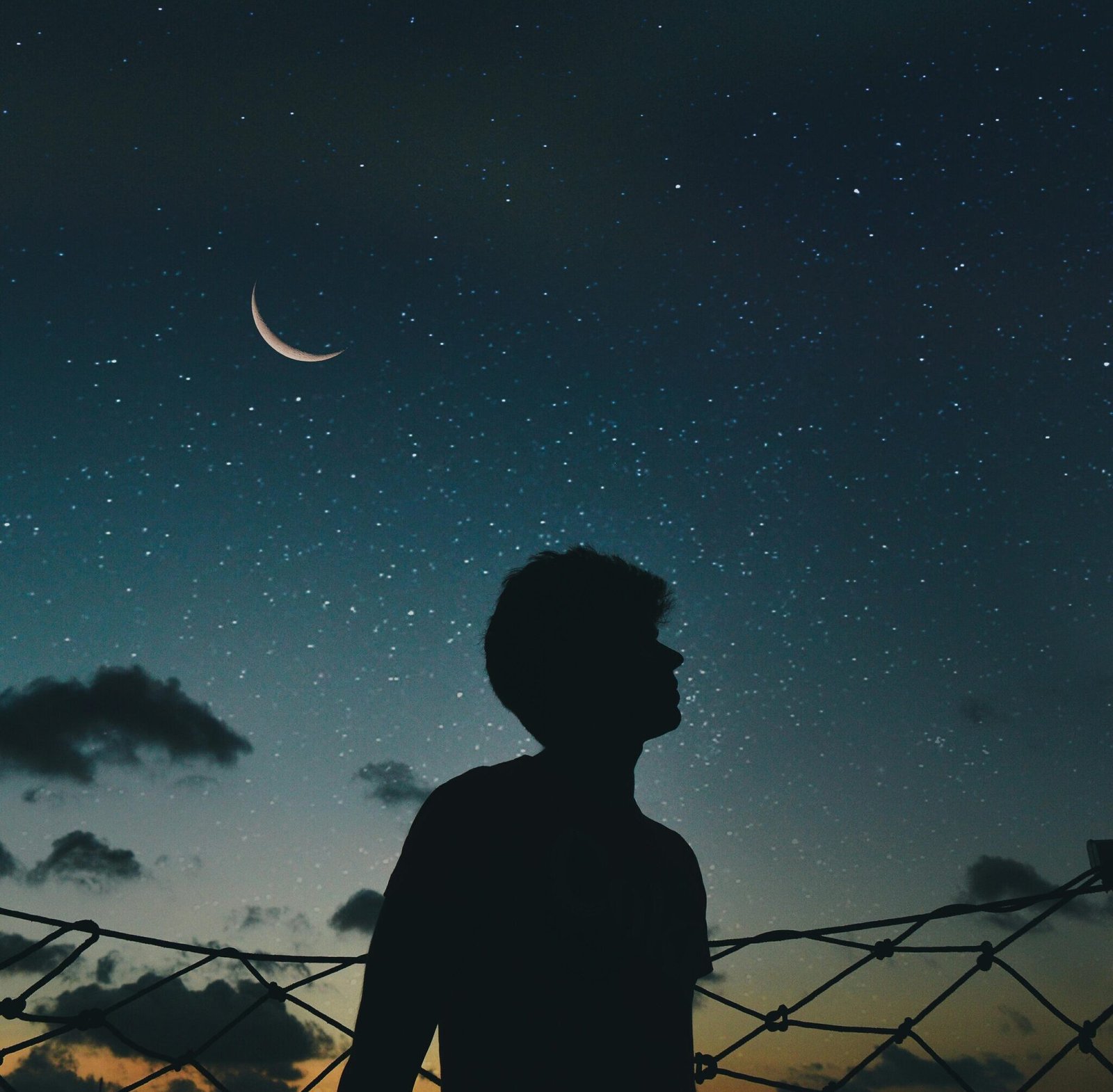 A lone silhouette stands beneath a crescent moon and starry sky, evoking a sense of solitude and wonder.