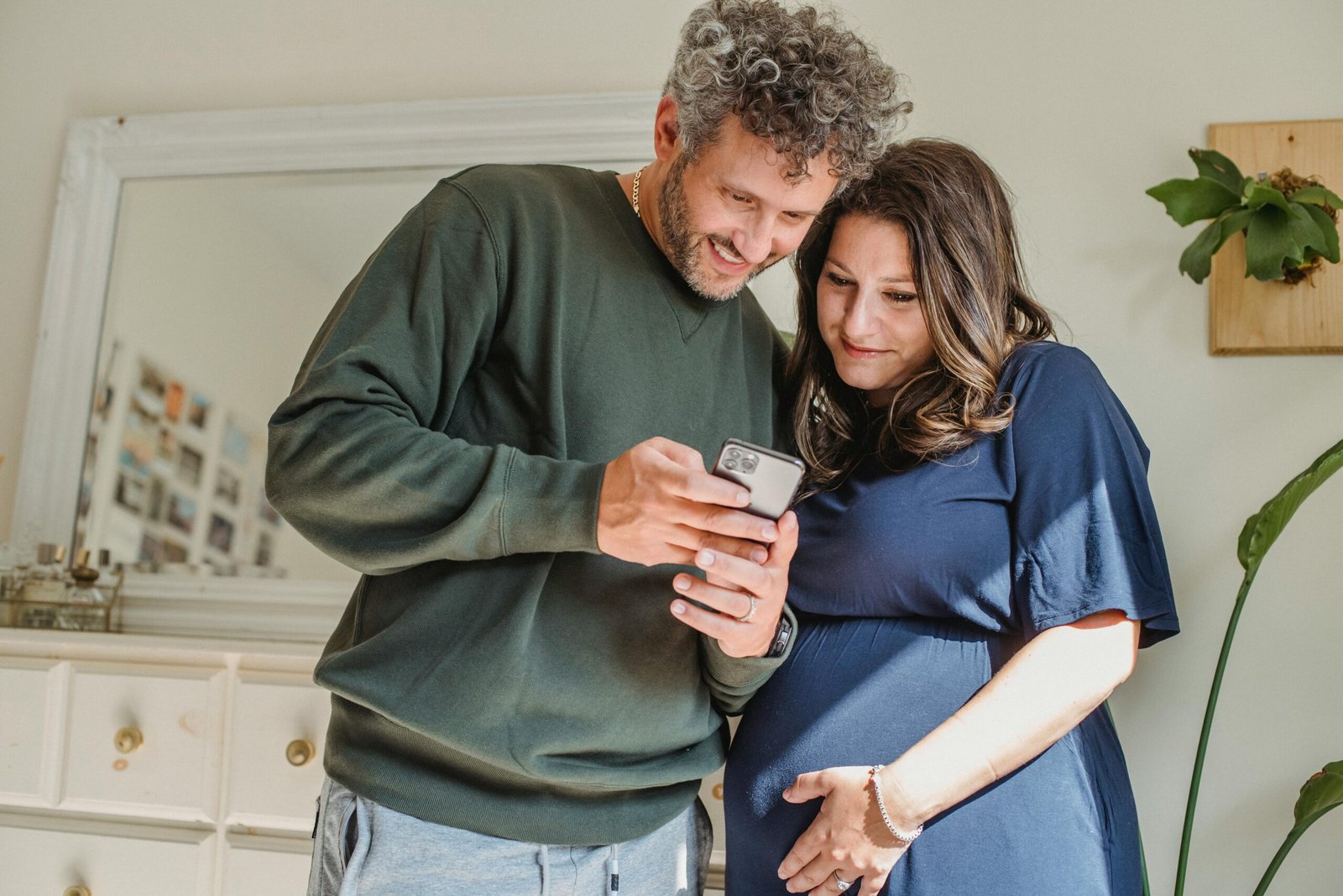 Content bearded male showing cellphone to feminine expectant woman caressing tummy in house room in sunlight