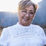Portrait of a smiling senior woman wearing glasses and a white blouse in natural sunlight.